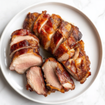 A bacon-wrapped pork tenderloin in a roasting pan, seasoned with garlic, brown sugar, and rosemary, ready to be roasted in the oven.