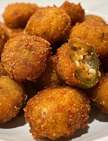 A plate of golden, crispy jalapeño popper bites piled high, with a side of creamy ranch dressing. The crunchy coating glistens under the light, hinting at the gooey, cheesy filling inside.