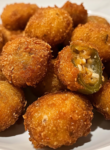 A plate of golden, crispy jalapeño popper bites piled high, with a side of creamy ranch dressing. The crunchy coating glistens under the light, hinting at the gooey, cheesy filling inside.